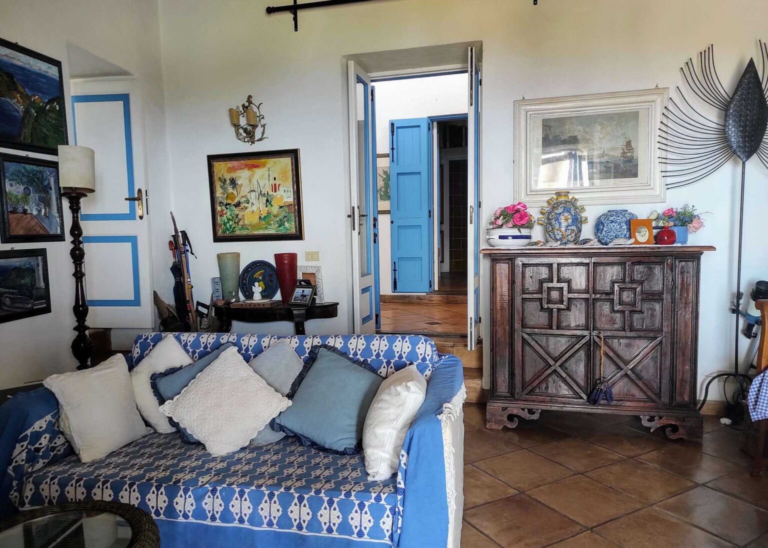 Cozy living room interior featuring a blue sofa with decorative pillows, a wooden cabinet with intricate designs, and vibrant artwork on the walls. A doorway with blue shutters leads outside, enhancing the bright and welcoming atmosphere. | Only on SopranoVillas