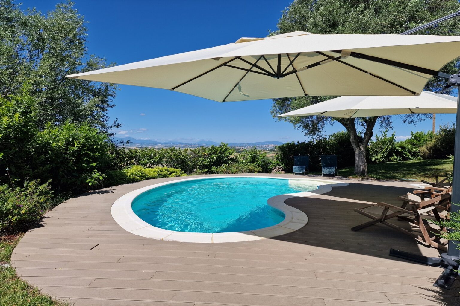 Luxury outdoor swimming pool with a curved design, surrounded by lush greenery and shaded by large umbrellas, offering a relaxing atmosphere against a clear blue sky. | Only on SopranoVillas