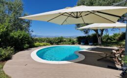 Luxury outdoor swimming pool with a curved design, surrounded by lush greenery and shaded by large umbrellas, offering a relaxing atmosphere against a clear blue sky.