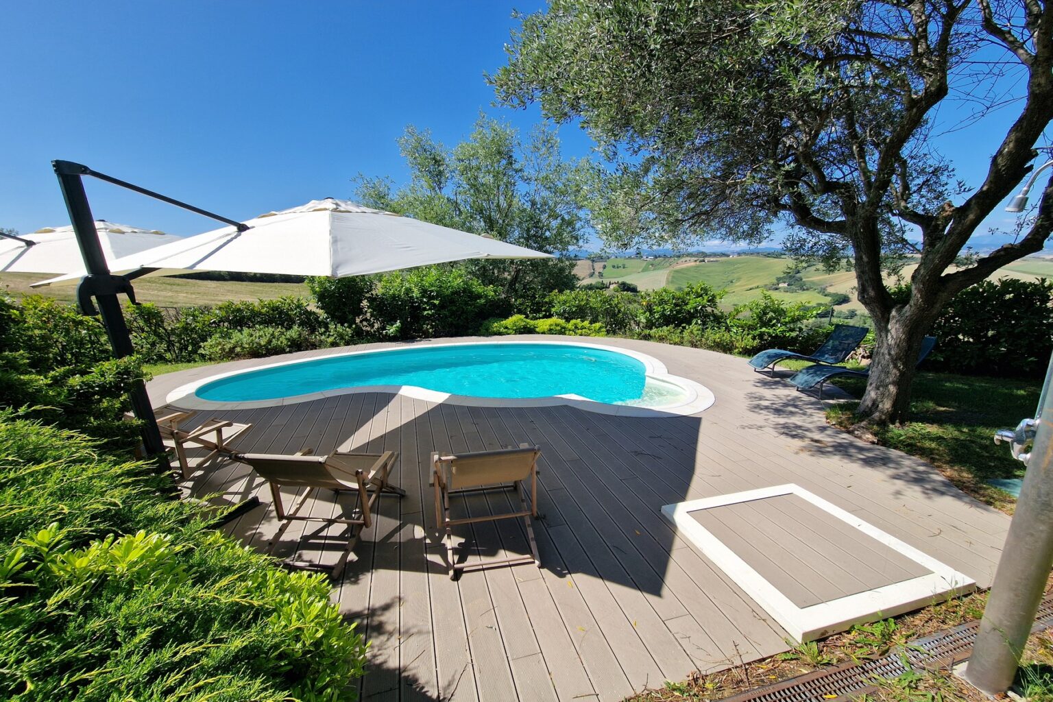 Scenic outdoor pool area with lounge chairs, umbrellas, and lush greenery overlooking rolling hills under a clear blue sky. Perfect for relaxation and enjoying nature. | Only on SopranoVillas