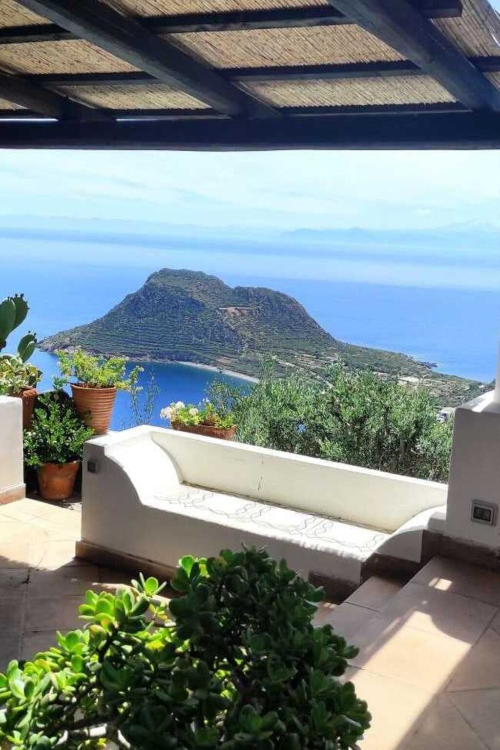 Stunning coastal view of a mountainous island surrounded by clear blue waters, framed by lush greenery and terracotta pots on a terrace. Ideal for travel and vacation planning. | Only on SopranoVillas