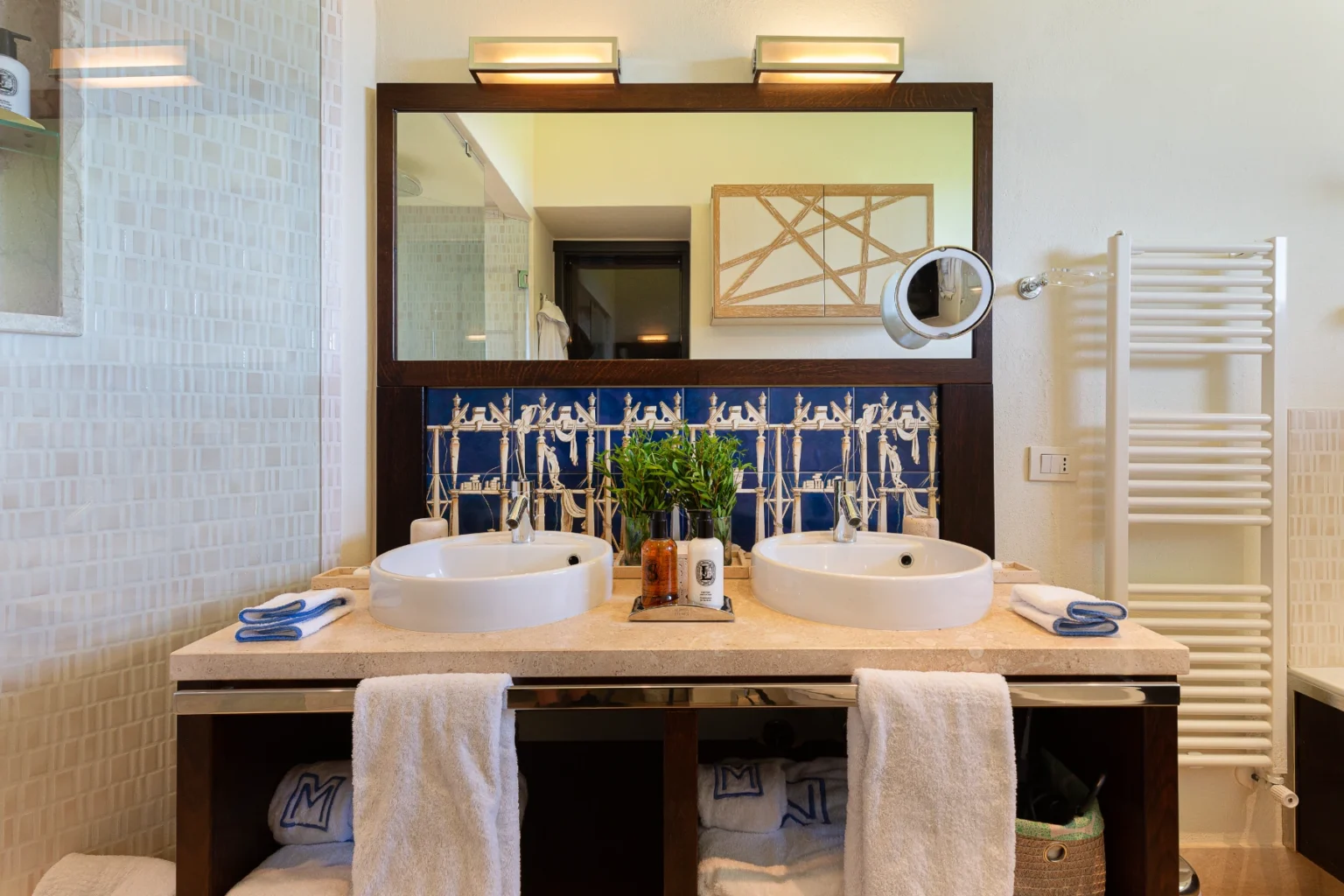 Modern bathroom featuring double sinks, a large mirror, decorative blue tile backsplash, and neatly arranged towels. The space includes stylish fixtures and a plant for added decor, creating a clean and inviting atmosphere. | Only on SopranoVillas