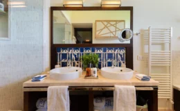 Modern bathroom featuring double sinks, a large mirror, decorative blue tile backsplash, and neatly arranged towels. The space includes stylish fixtures and a plant for added decor, creating a clean and inviting atmosphere.