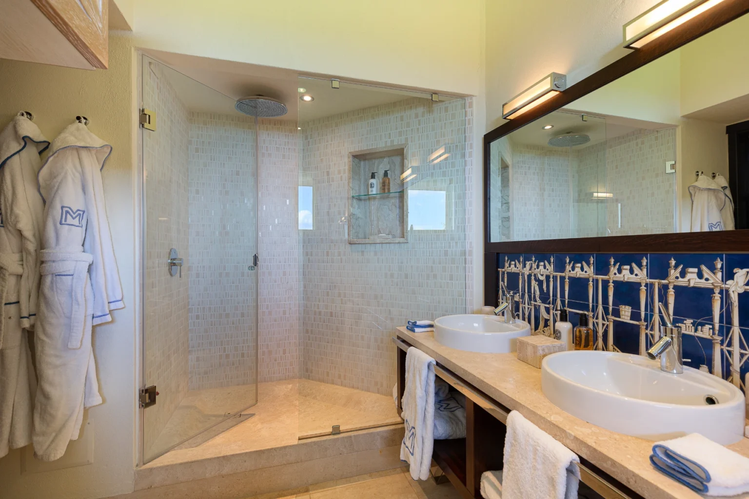 Modern bathroom featuring a spacious walk-in shower with glass enclosure, dual sinks, and elegant stone countertops. Fluffy white bathrobes hang nearby, complemented by stylish blue tile accents. Ideal for a luxurious home or hotel setting. | Only on SopranoVillas