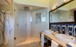 Modern bathroom featuring a spacious walk-in shower with glass enclosure, dual sinks, and elegant stone countertops. Fluffy white bathrobes hang nearby, complemented by stylish blue tile accents. Ideal for a luxurious home or hotel setting.