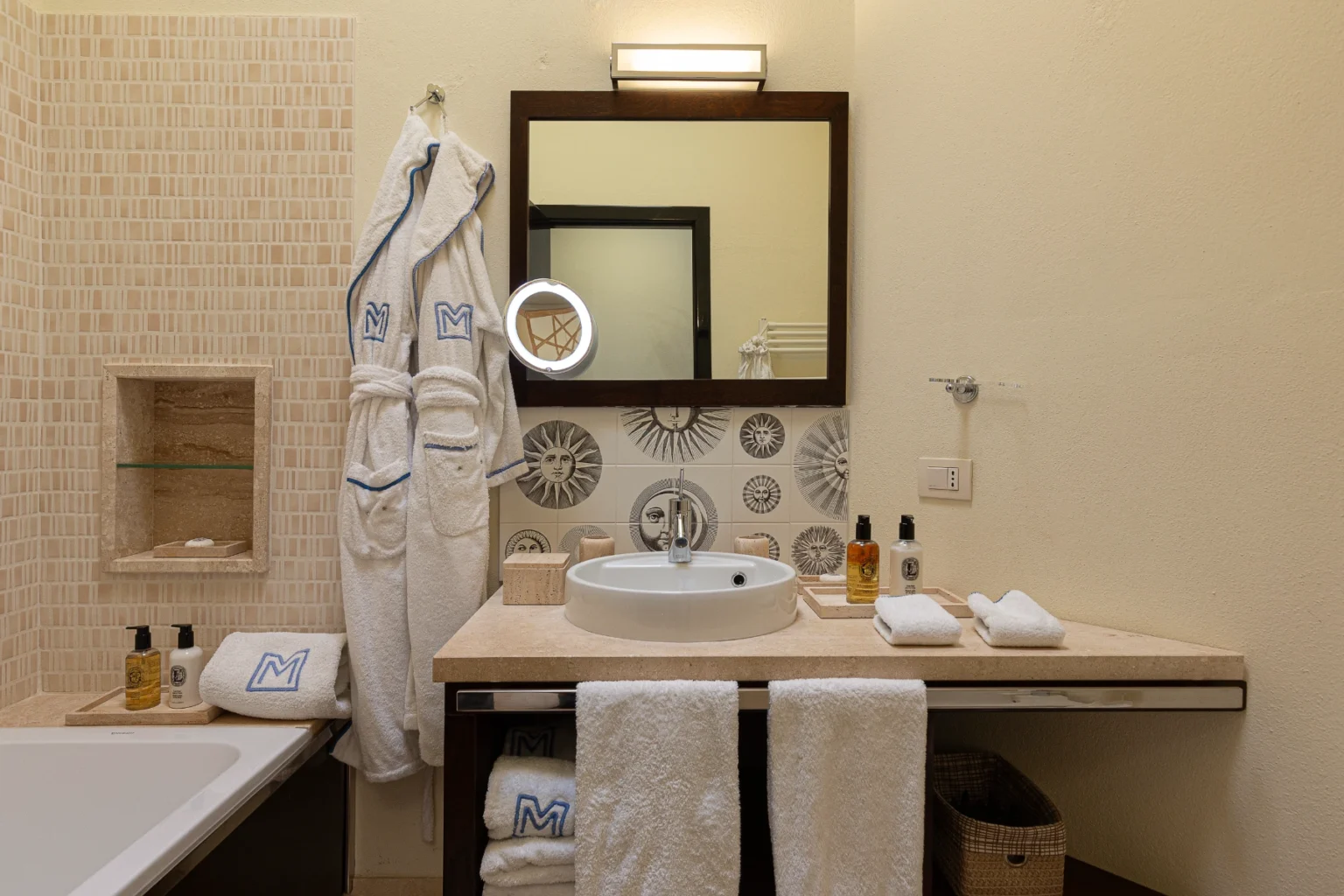 Modern bathroom interior featuring a stylish sink, decorative tiles with sun motifs, plush white bathrobes, and high-quality toiletries. The space is illuminated by a sleek mirror light, providing a luxurious and inviting atmosphere. | Only on SopranoVillas