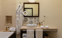 Modern bathroom interior featuring a stylish sink, decorative tiles with sun motifs, plush white bathrobes, and high-quality toiletries. The space is illuminated by a sleek mirror light, providing a luxurious and inviting atmosphere.