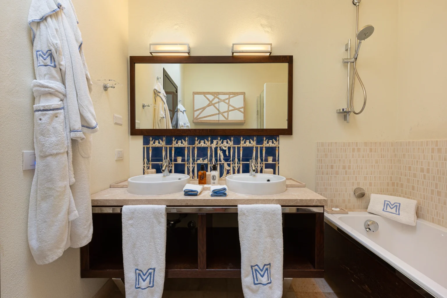 Luxurious hotel bathroom featuring double sinks, a large mirror, and elegant decor. Includes a bathtub, plush white robes, and towels with a monogram. Modern fixtures and stylish blue tile accents create a relaxing atmosphere. | Only on SopranoVillas