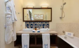 Luxurious hotel bathroom featuring double sinks, a large mirror, and elegant decor. Includes a bathtub, plush white robes, and towels with a monogram. Modern fixtures and stylish blue tile accents create a relaxing atmosphere.
