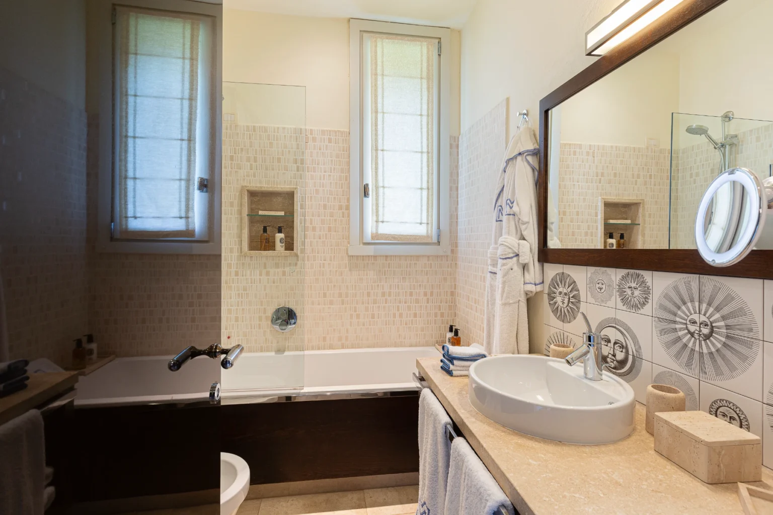 Modern bathroom featuring a bathtub with glass screen, a stylish sink with a mirror, and decorative wall tiles. The space includes natural light from a window and neatly arranged toiletries, emphasizing a clean and inviting atmosphere. | Only on SopranoVillas