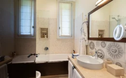 Modern bathroom featuring a bathtub with glass screen, a stylish sink with a mirror, and decorative wall tiles. The space includes natural light from a window and neatly arranged toiletries, emphasizing a clean and inviting atmosphere.