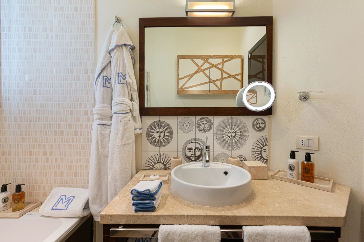 Stylish bathroom featuring a modern sink, decorative sun-themed tiles, plush white bathrobes, neatly arranged towels, and luxury toiletries on a marble countertop, complemented by a large mirror and warm lighting. | Only on SopranoVillas
