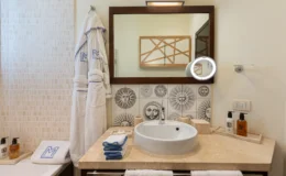 Stylish bathroom featuring a modern sink, decorative sun-themed tiles, plush white bathrobes, neatly arranged towels, and luxury toiletries on a marble countertop, complemented by a large mirror and warm lighting.