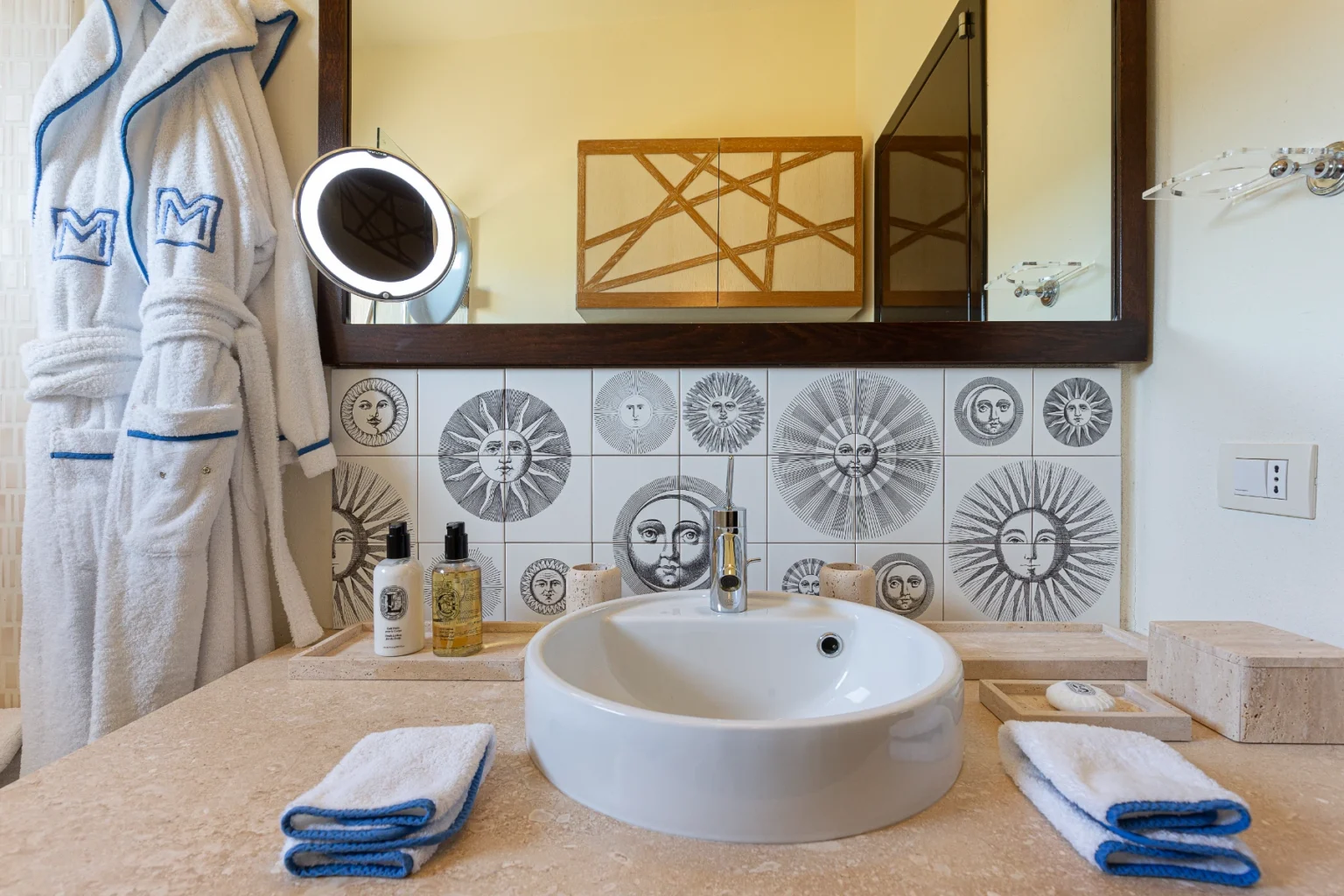 Stylish bathroom featuring a round white sink, decorative sun-patterned tile backsplash, a large mirror with a backlight, and neatly arranged toiletries. A plush white robe hangs beside the sink, complemented by folded towels with blue trim on a beige countertop. | Only on SopranoVillas