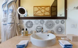 Stylish bathroom featuring a round white sink, decorative sun-patterned tile backsplash, a large mirror with a backlight, and neatly arranged toiletries. A plush white robe hangs beside the sink, complemented by folded towels with blue trim on a beige countertop.