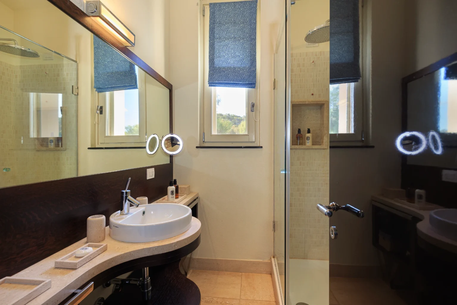 Modern bathroom interior featuring a curved countertop with a round sink, a large mirror with integrated lighting, and a glass shower. The room is well-lit with natural light from a window adorned with a blue fabric shade, showcasing a clean and stylish design. | Only on SopranoVillas