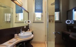 Modern bathroom interior featuring a curved countertop with a round sink, a large mirror with integrated lighting, and a glass shower. The room is well-lit with natural light from a window adorned with a blue fabric shade, showcasing a clean and stylish design.