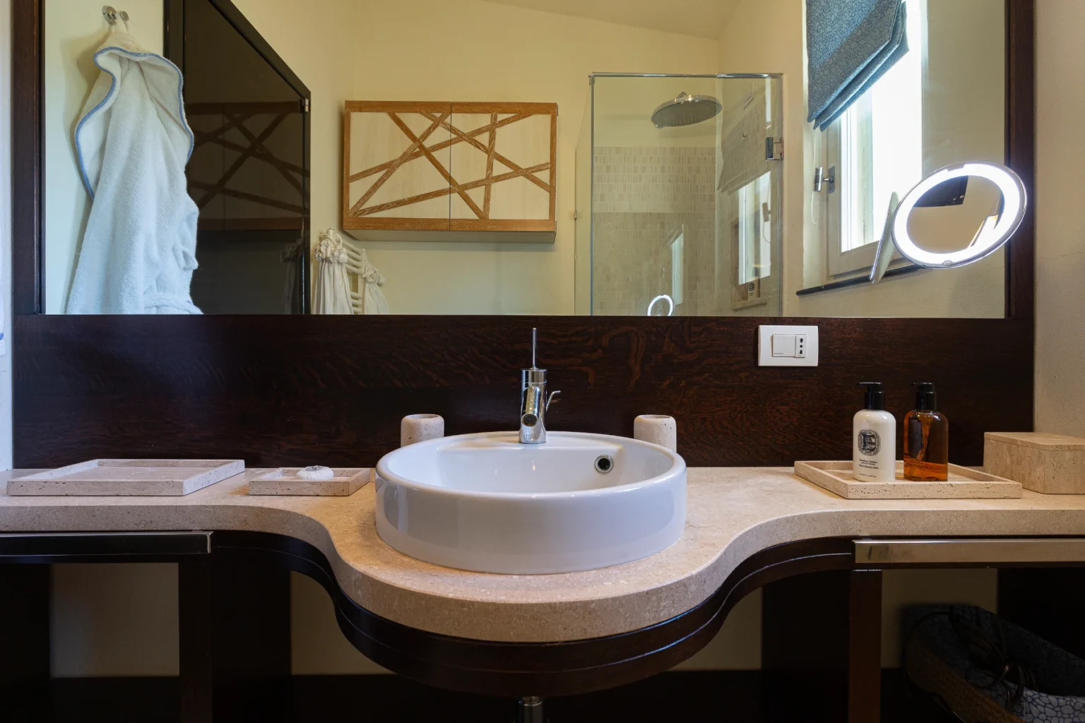 Modern bathroom interior featuring a round white sink on a curved stone countertop, wooden cabinetry with geometric design, a glass shower enclosure, and a stylish illuminated mirror. Decorative elements include a soft blue towel and personal care products in sleek bottles. | Only on SopranoVillas