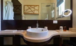Modern bathroom interior featuring a round white sink on a curved stone countertop, wooden cabinetry with geometric design, a glass shower enclosure, and a stylish illuminated mirror. Decorative elements include a soft blue towel and personal care products in sleek bottles.