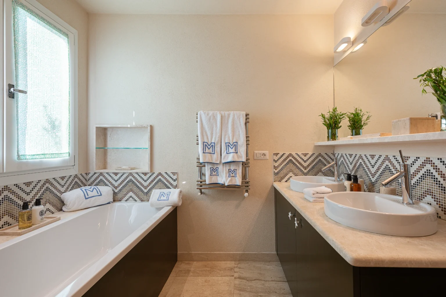 Modern bathroom interior featuring a freestanding bathtub, double sinks, and stylish mosaic tiles. Soft lighting highlights the elegant design, complemented by plush towels and decorative plants. The space exudes a clean and contemporary aesthetic. | Only on SopranoVillas