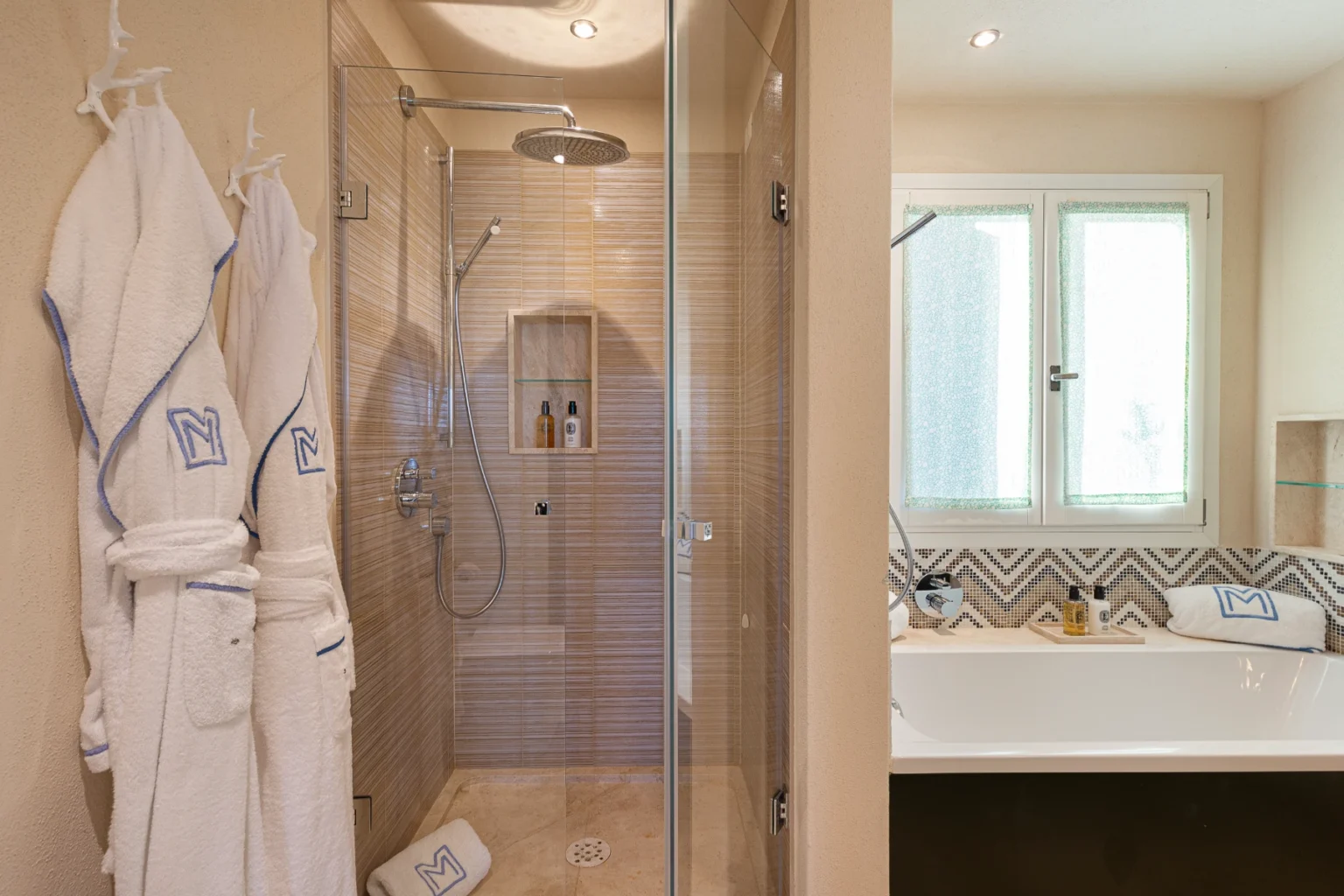 Modern bathroom featuring a glass-enclosed shower with a rainfall showerhead, beige tiled walls, and a built-in shelf for toiletries. Plush white bathrobes hang on hooks, while a freestanding bathtub is visible in the corner, accented by patterned tiles. Natural light filters through frosted windows, creating a serene atmosphere. | Only on SopranoVillas
