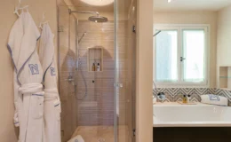 Modern bathroom featuring a glass-enclosed shower with a rainfall showerhead, beige tiled walls, and a built-in shelf for toiletries. Plush white bathrobes hang on hooks, while a freestanding bathtub is visible in the corner, accented by patterned tiles. Natural light filters through frosted windows, creating a serene atmosphere.