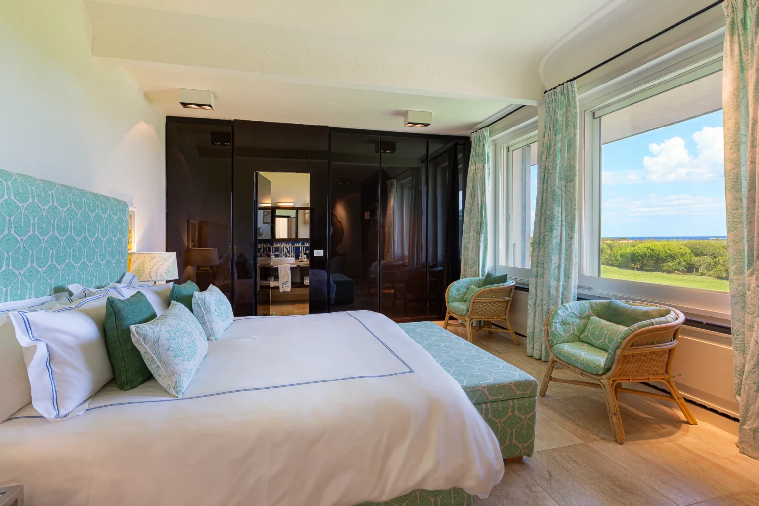 Elegant hotel room featuring a plush king-size bed with decorative pillows, modern furnishings, and large windows showcasing a scenic view. The room includes stylish rattan chairs, soft green accents, and a sleek black wardrobe, creating a serene and inviting atmosphere. Ideal for a relaxing getaway. | Only on SopranoVillas