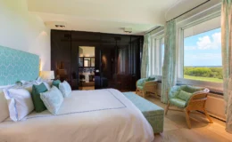 Elegant hotel room featuring a plush king-size bed with decorative pillows, modern furnishings, and large windows showcasing a scenic view. The room includes stylish rattan chairs, soft green accents, and a sleek black wardrobe, creating a serene and inviting atmosphere. Ideal for a relaxing getaway.