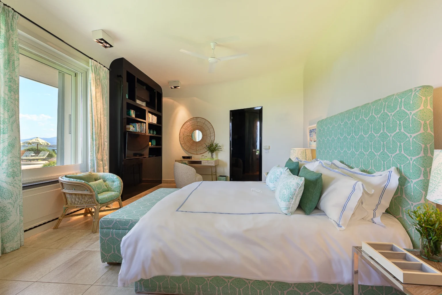 Modern bedroom design featuring a king-sized bed with a patterned headboard, decorative pillows, and stylish bedside lamps. The room includes a cozy seating area with a wicker chair, a sleek black entertainment unit, and a large window offering scenic views. Natural light enhances the serene atmosphere, complemented by soft color tones and elegant decor. | Only on SopranoVillas