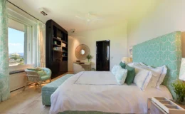 Modern bedroom design featuring a king-sized bed with a patterned headboard, decorative pillows, and stylish bedside lamps. The room includes a cozy seating area with a wicker chair, a sleek black entertainment unit, and a large window offering scenic views. Natural light enhances the serene atmosphere, complemented by soft color tones and elegant decor.