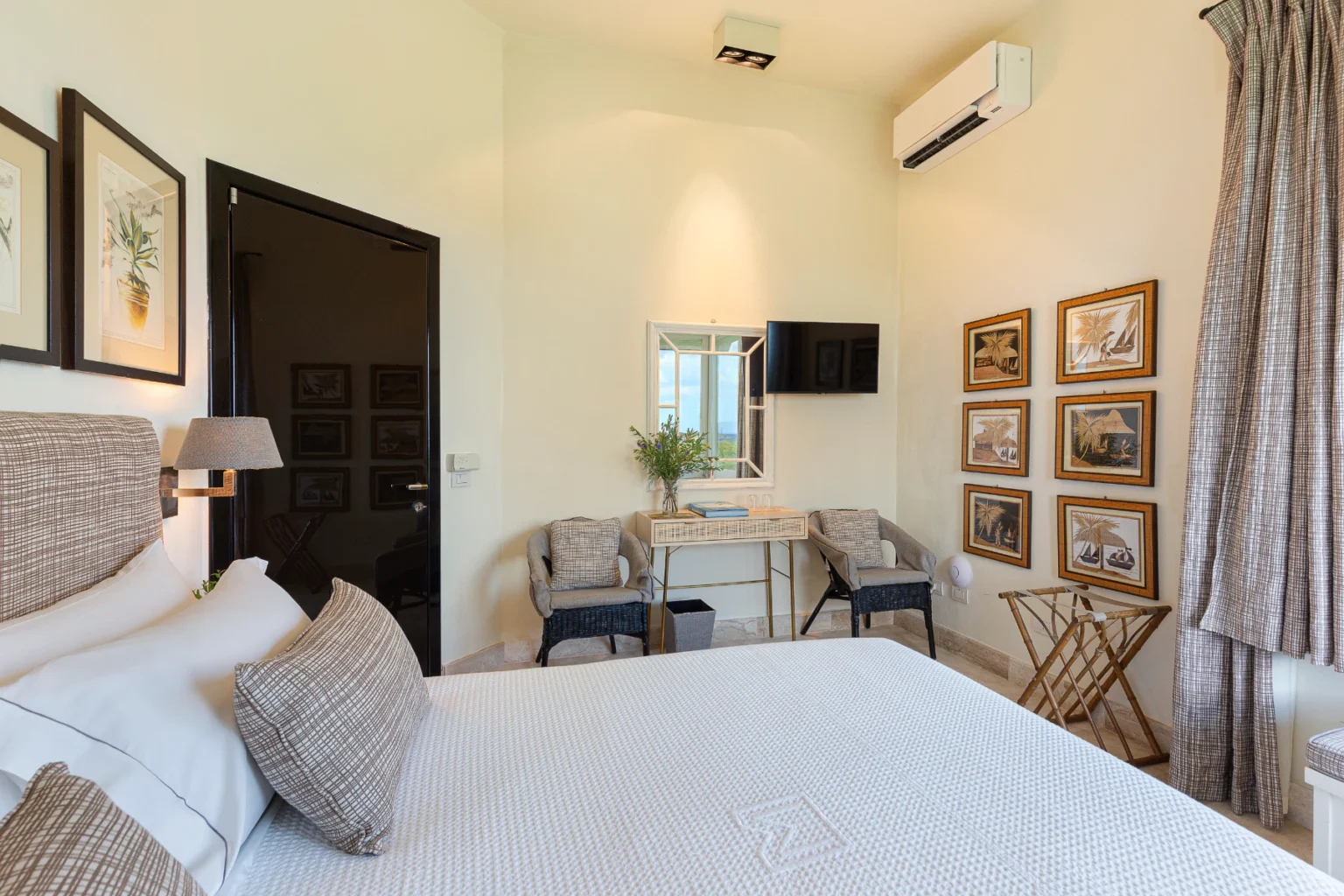 Cozy bedroom featuring a plush bed with decorative pillows, modern air conditioning unit, wall-mounted TV, and stylish seating area. The room is adorned with framed artwork and a mirror, creating a serene and inviting atmosphere. | Only on SopranoVillas
