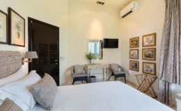 Cozy bedroom featuring a plush bed with decorative pillows, modern air conditioning unit, wall-mounted TV, and stylish seating area. The room is adorned with framed artwork and a mirror, creating a serene and inviting atmosphere.