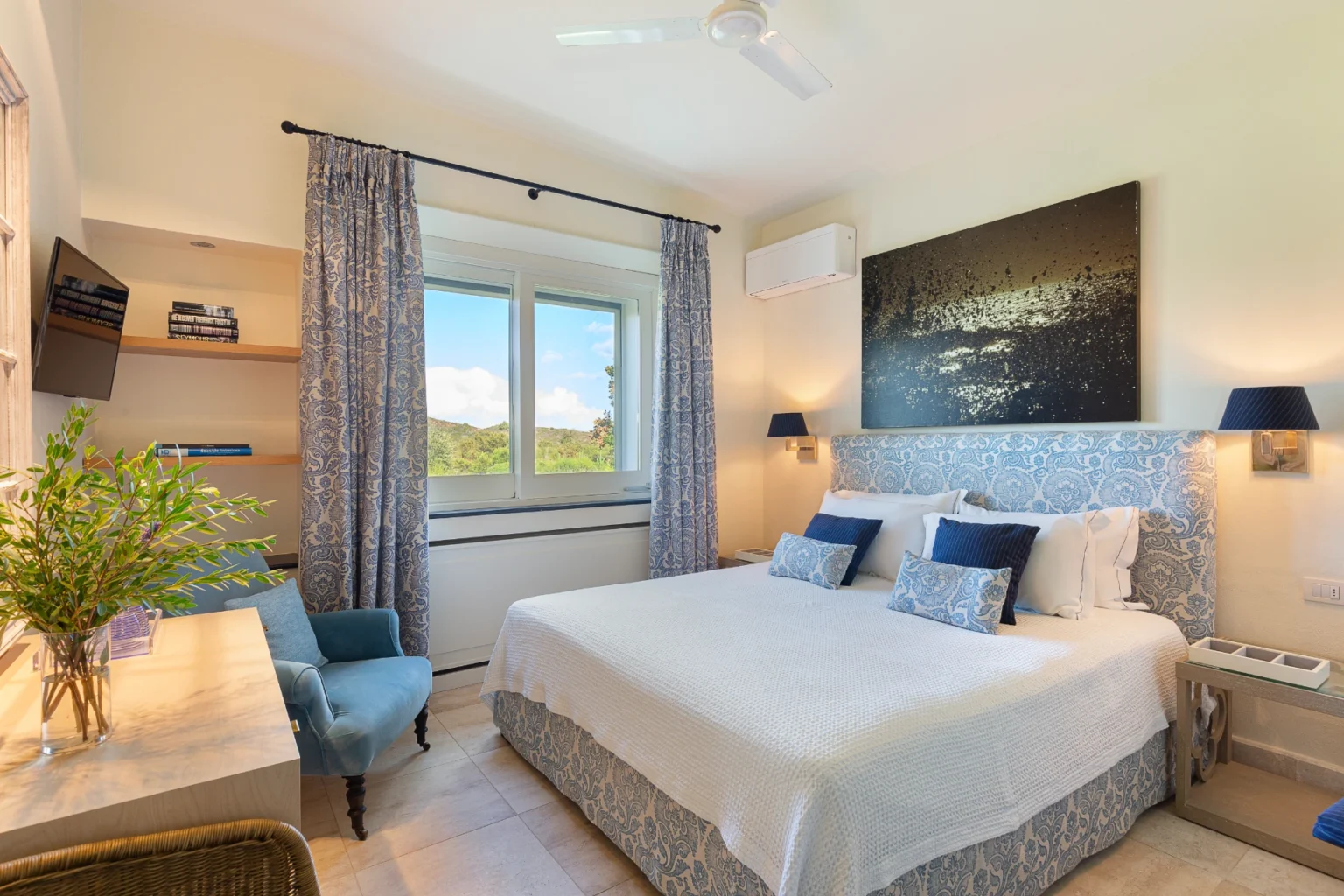 Bright and airy bedroom featuring a queen-sized bed with blue and white patterned bedding, elegant curtains, and a cozy reading chair. The room includes a small desk, a flat-screen TV, and large windows showcasing scenic views, creating a tranquil and inviting atmosphere. Perfect for relaxation and comfort. | Only on SopranoVillas