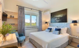 Bright and airy bedroom featuring a queen-sized bed with blue and white patterned bedding, elegant curtains, and a cozy reading chair. The room includes a small desk, a flat-screen TV, and large windows showcasing scenic views, creating a tranquil and inviting atmosphere. Perfect for relaxation and comfort.