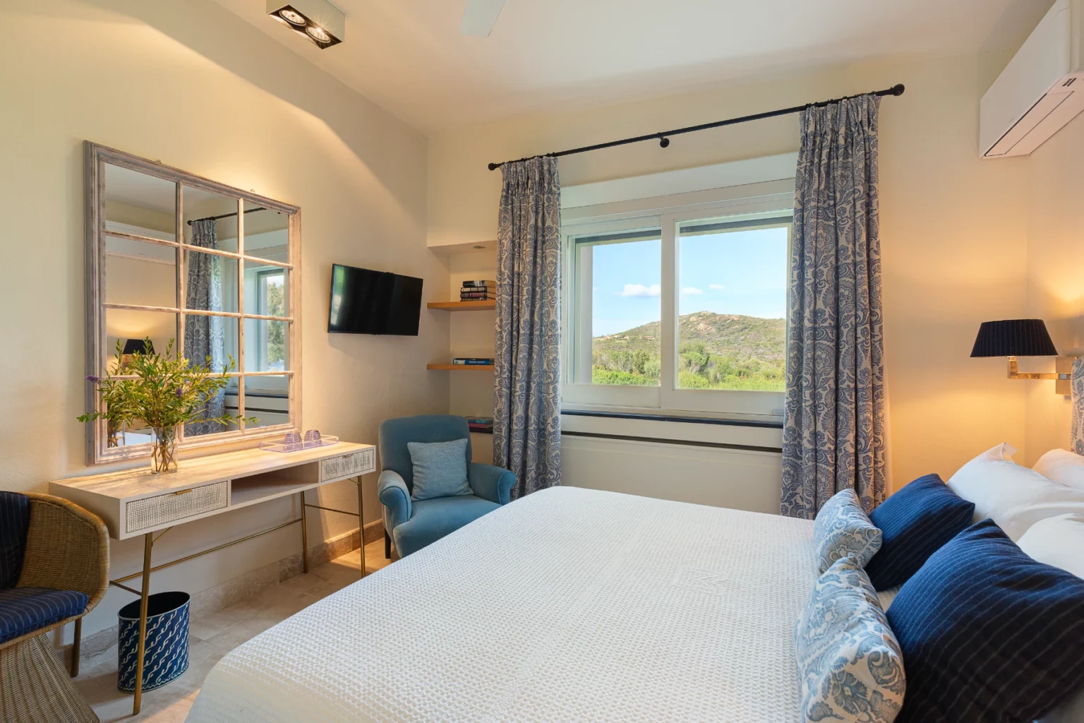 Bright and airy bedroom featuring a comfortable bed with decorative pillows, a stylish desk with a vase of greenery, and a cozy armchair. Large window offers a scenic view of hills, while elegant curtains and modern lighting enhance the decor. A flat-screen TV is mounted on the wall, and books are neatly arranged on a shelf. | Only on SopranoVillas