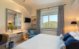 Bright and airy bedroom featuring a comfortable bed with decorative pillows, a stylish desk with a vase of greenery, and a cozy armchair. Large window offers a scenic view of hills, while elegant curtains and modern lighting enhance the decor. A flat-screen TV is mounted on the wall, and books are neatly arranged on a shelf.