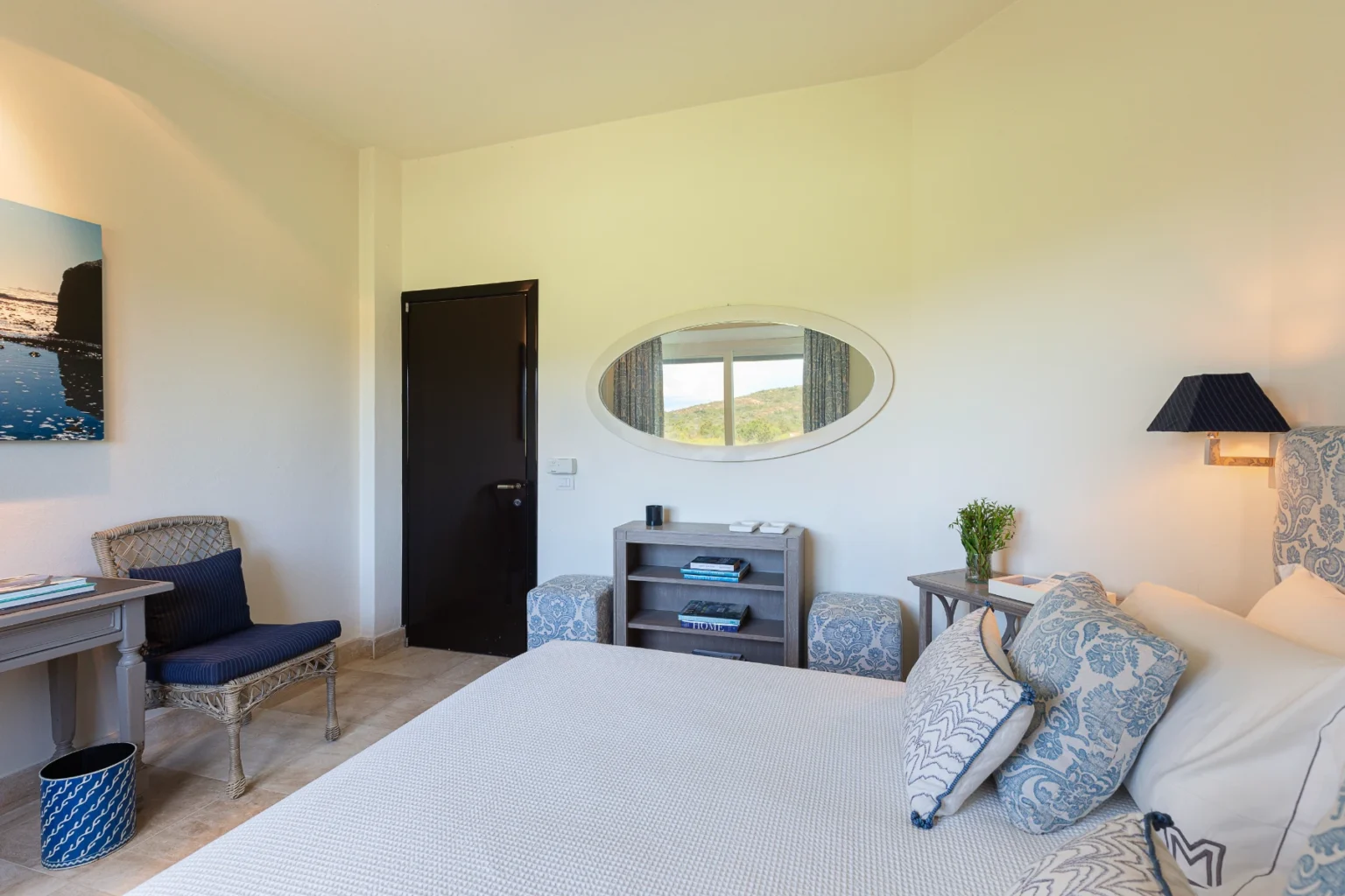 Cozy hotel room featuring a comfortable bed with decorative pillows, a stylish desk with a chair, and natural light from a window with scenic views. The room includes a modern lamp, a storage shelf with books, and a touch of greenery. Ideal for relaxation and comfort during a getaway. | Only on SopranoVillas