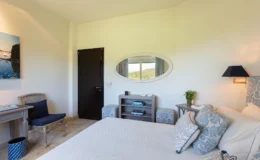 Cozy hotel room featuring a comfortable bed with decorative pillows, a stylish desk with a chair, and natural light from a window with scenic views. The room includes a modern lamp, a storage shelf with books, and a touch of greenery. Ideal for relaxation and comfort during a getaway.