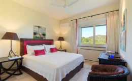 Cozy hotel bedroom featuring a king-sized bed with decorative pillows, bedside lamps, and a scenic window view. The room is adorned with light-colored walls, a ceiling fan, and stylish furniture, creating a relaxing atmosphere.