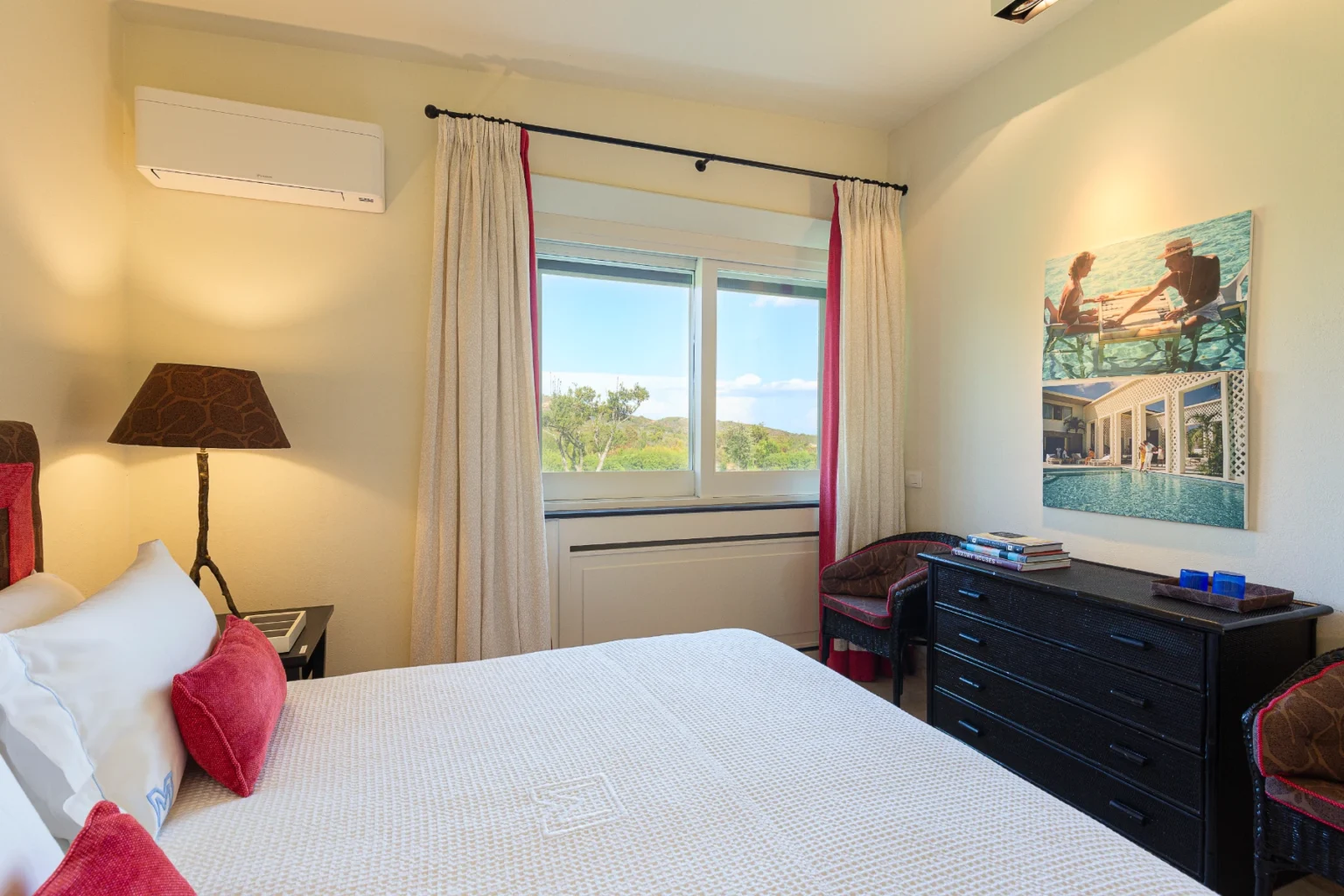 Cozy bedroom featuring a comfortable bed with decorative pillows, a stylish lamp, and a dresser. Large window provides a view of greenery, while curtains add a touch of elegance. Wall art depicts leisure activities by a pool, enhancing the room's inviting atmosphere. | Only on SopranoVillas