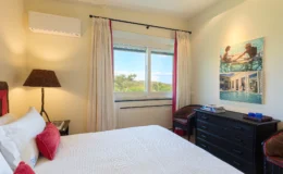 Cozy bedroom featuring a comfortable bed with decorative pillows, a stylish lamp, and a dresser. Large window provides a view of greenery, while curtains add a touch of elegance. Wall art depicts leisure activities by a pool, enhancing the room's inviting atmosphere.