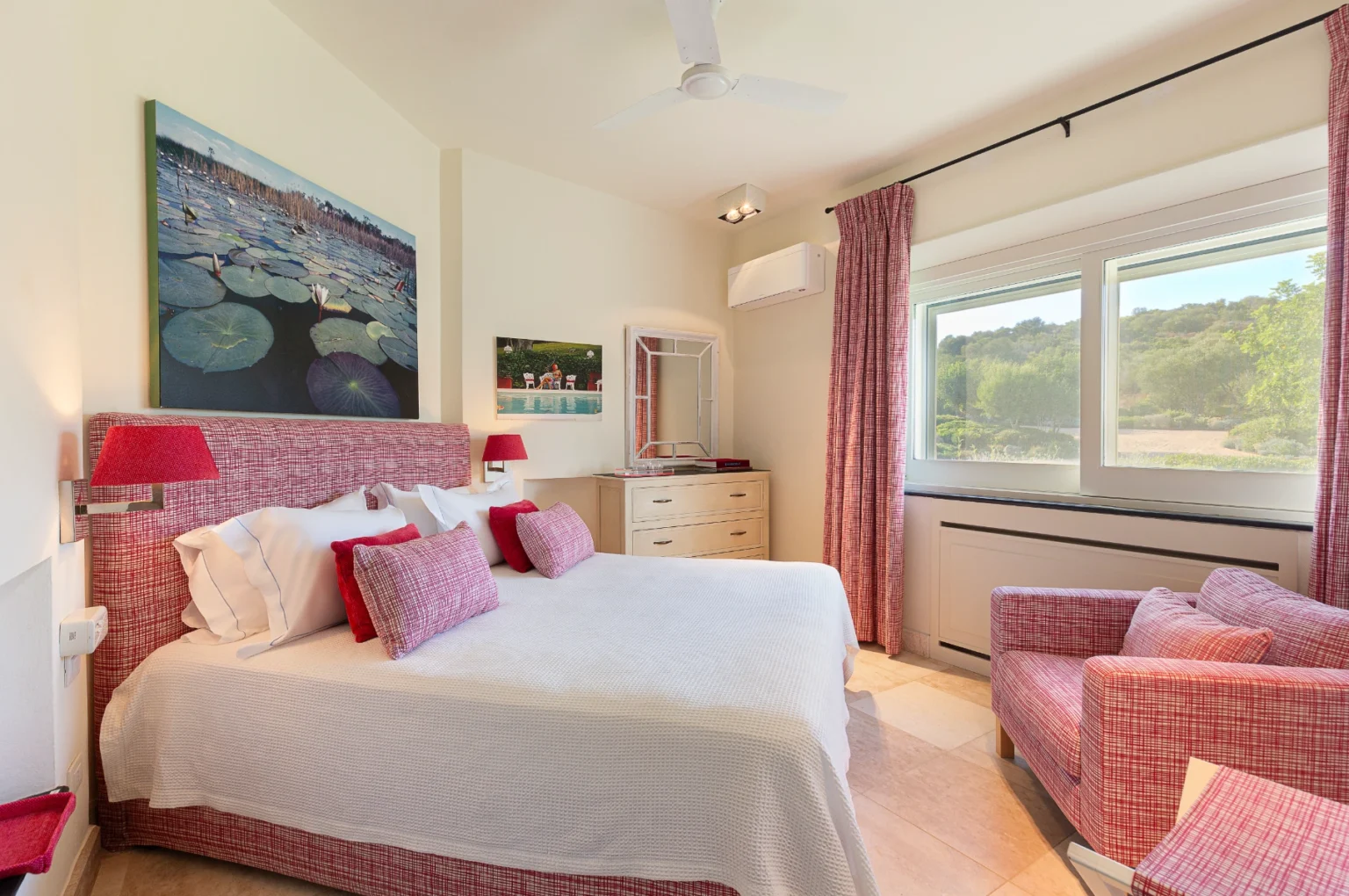 Cozy bedroom featuring a queen-sized bed with red and white patterned bedding, complemented by decorative pillows. The room includes a comfortable armchair, a wooden dresser, and artwork depicting a serene water lily scene. Natural light floods in through large windows, offering a view of lush greenery outside. Stylish curtains and modern lighting enhance the inviting atmosphere. | Only on SopranoVillas