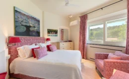 Cozy bedroom featuring a queen-sized bed with red and white patterned bedding, complemented by decorative pillows. The room includes a comfortable armchair, a wooden dresser, and artwork depicting a serene water lily scene. Natural light floods in through large windows, offering a view of lush greenery outside. Stylish curtains and modern lighting enhance the inviting atmosphere.