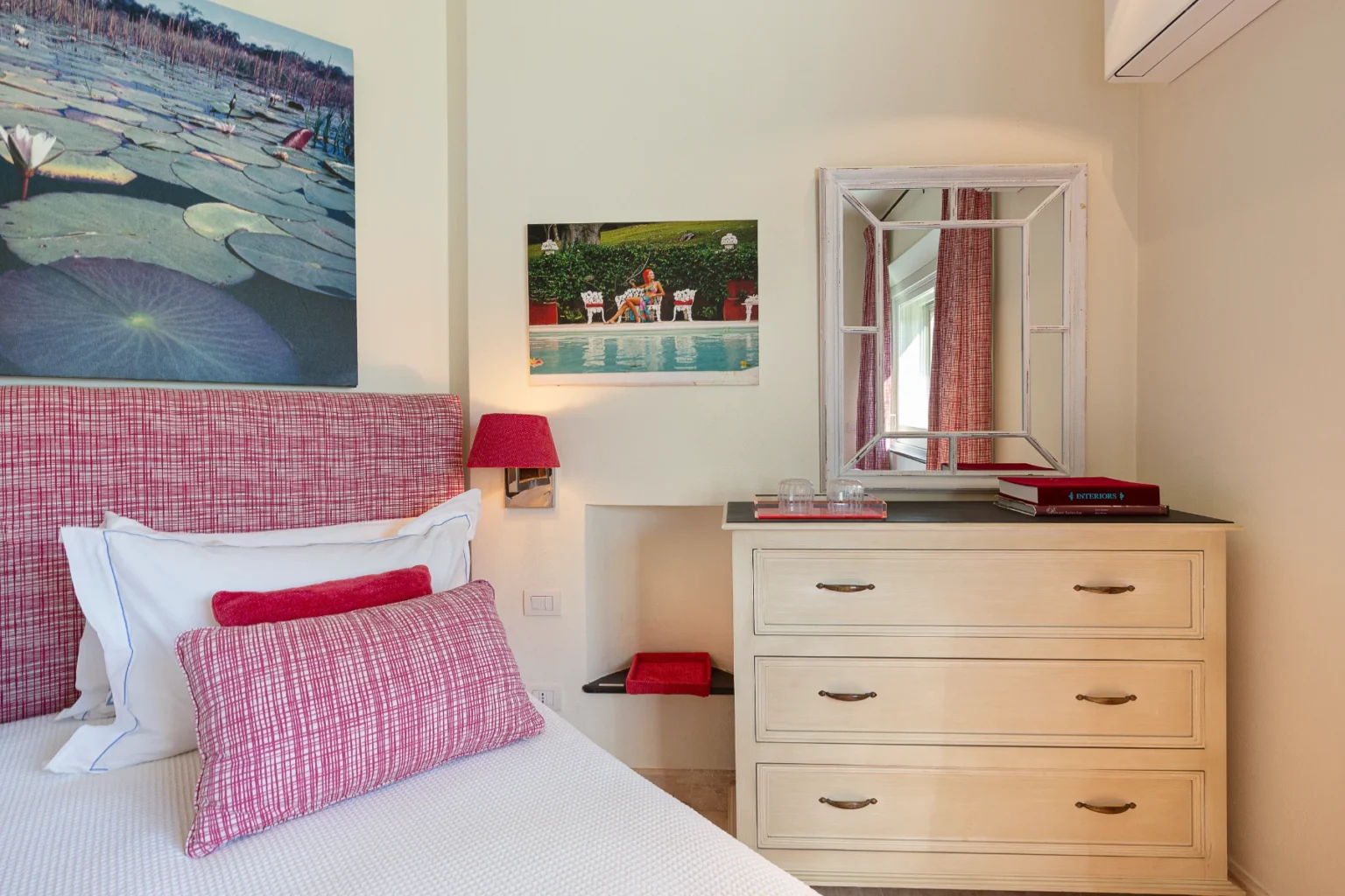 Cozy bedroom featuring a pink patterned headboard with decorative pillows, a light wooden dresser, and a stylish mirror. Artwork of a tranquil lily pond and a vibrant pool scene adorn the walls, complemented by a bedside lamp and decorative accents. Ideal for a relaxing retreat. | Only on SopranoVillas