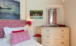 Cozy bedroom featuring a pink patterned headboard with decorative pillows, a light wooden dresser, and a stylish mirror. Artwork of a tranquil lily pond and a vibrant pool scene adorn the walls, complemented by a bedside lamp and decorative accents. Ideal for a relaxing retreat.