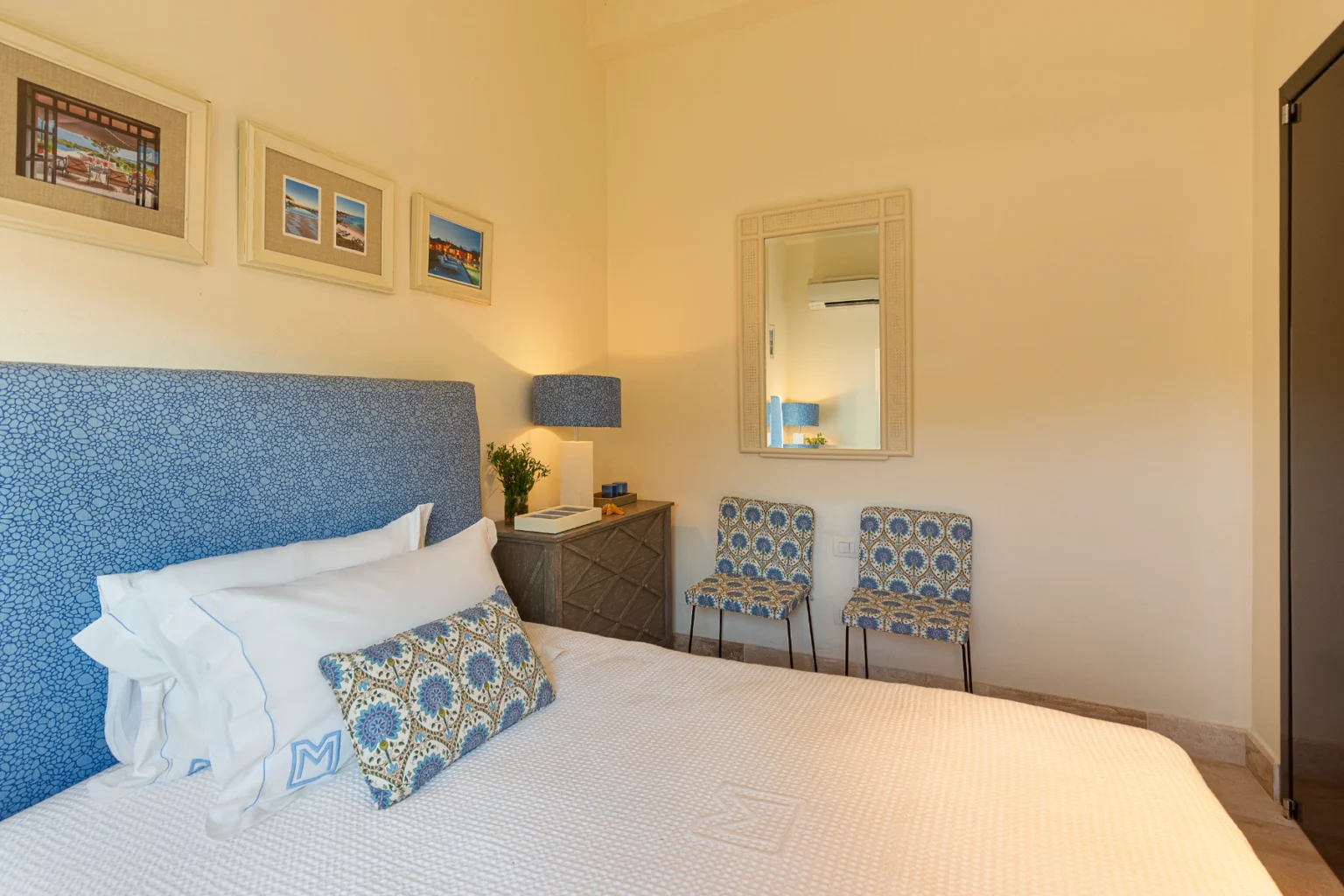 Cozy bedroom featuring a blue patterned headboard, white bedding, decorative pillows, and framed coastal artwork on the walls. The room includes a stylish lamp, a small nightstand, and two patterned chairs, creating a welcoming and elegant atmosphere. | Only on SopranoVillas