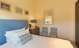 Cozy bedroom featuring a blue patterned headboard, white bedding, decorative pillows, and framed coastal artwork on the walls. The room includes a stylish lamp, a small nightstand, and two patterned chairs, creating a welcoming and elegant atmosphere.
