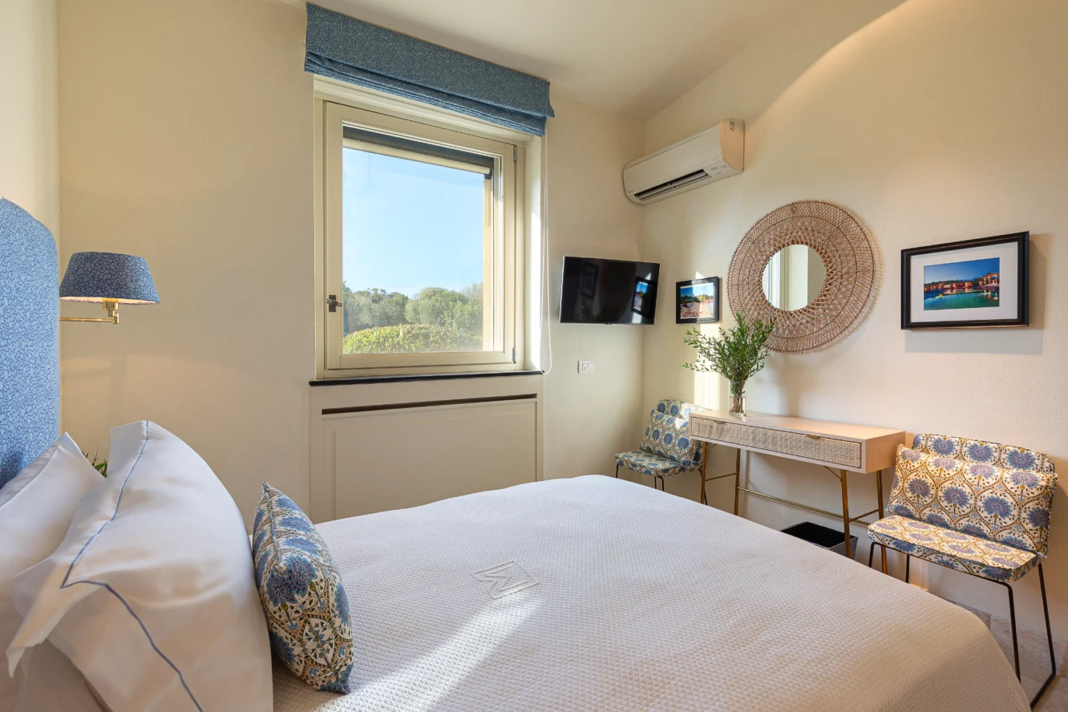 Cozy hotel bedroom featuring a comfortable bed with decorative pillows, stylish chairs, a modern TV, and a large window offering natural light and a view of greenery. The room is adorned with elegant decor, including a round mirror and a small plant on a desk, creating a welcoming atmosphere for guests. | Only on SopranoVillas