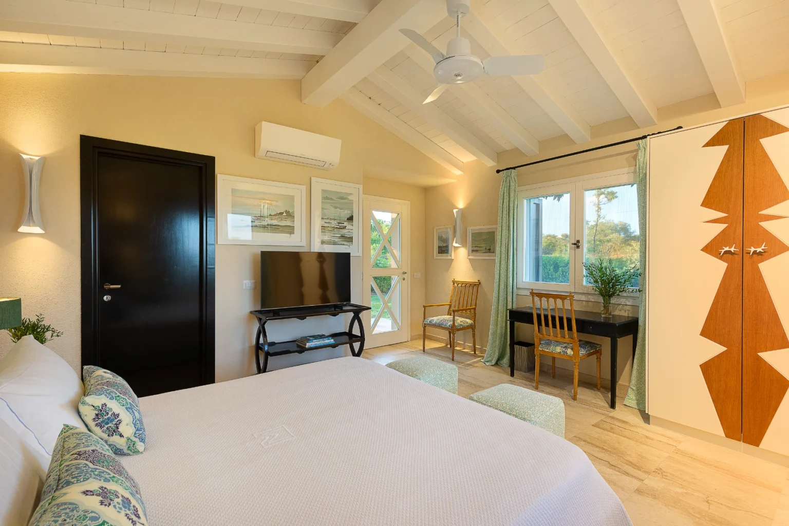Cozy bedroom interior featuring a queen-sized bed with decorative pillows, a modern TV stand, and a stylish workspace. The room has wooden beams, soft lighting, and large windows with green curtains that overlook a scenic view. A wardrobe with unique design accents complements the decor. Ideal for relaxation and comfort. | Only on SopranoVillas