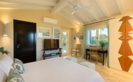 Cozy bedroom interior featuring a queen-sized bed with decorative pillows, a modern TV stand, and a stylish workspace. The room has wooden beams, soft lighting, and large windows with green curtains that overlook a scenic view. A wardrobe with unique design accents complements the decor. Ideal for relaxation and comfort.